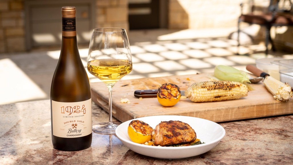 A bottle of 1924 Double Gold Buttery Chardonnay standing next to a chilled glass of wine on an outdoor grill styled tabletop.