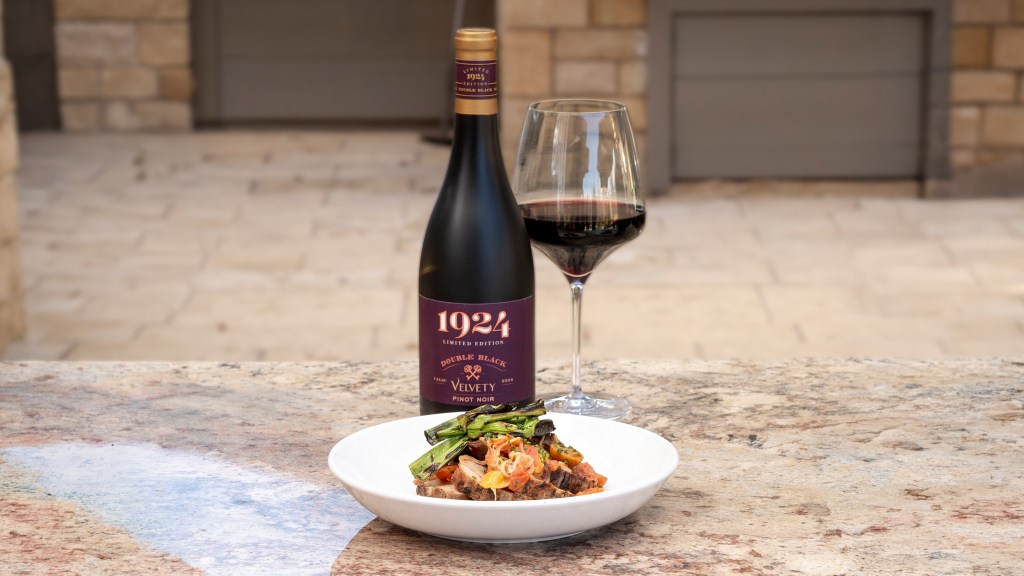 A bottle of 1924 Velvety Pinot Noir along with a glass of wine and food pairing on an outdoor tabletop.